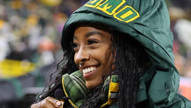 Simone Biles poses in a green jacket with her husband’s name stitched on the hood and smiles to the side.